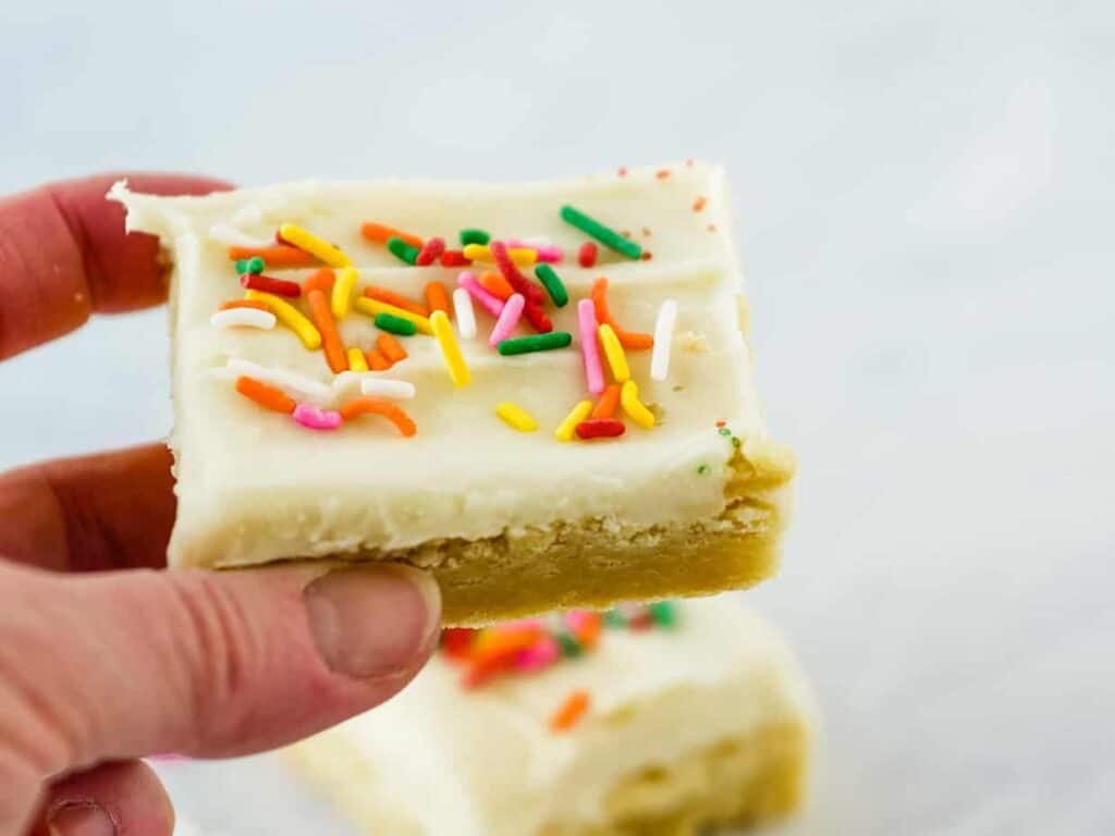 A hand holding a piece of vanilla frosted sugar cookie bar topped with rainbow sprinkles.