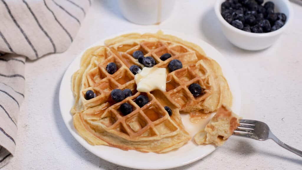 Belgian waffle topped with butter and syrup, garnished with blueberries, accompanied by a bowl of extra blueberries.