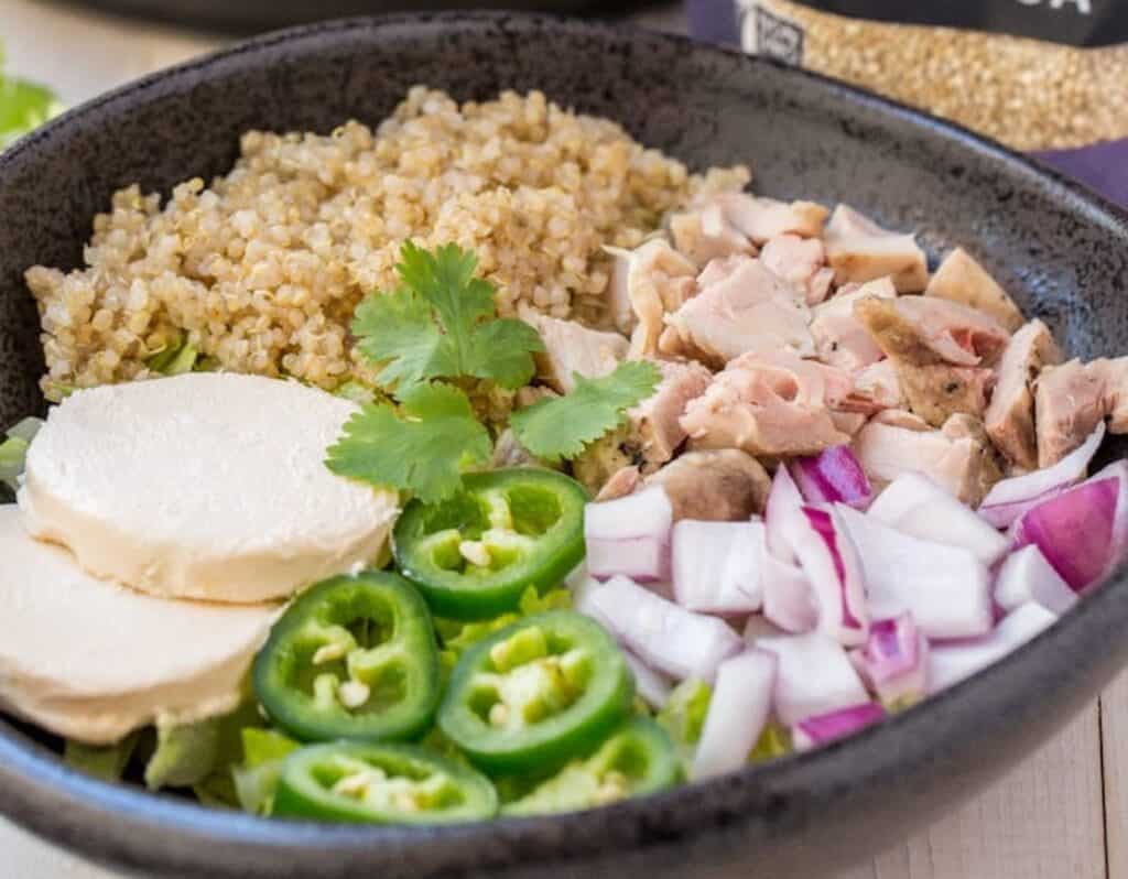 A bowl containing quinoa, sliced chicken, mozzarella cheese, red onions, cilantro, and sliced jalapeños.
