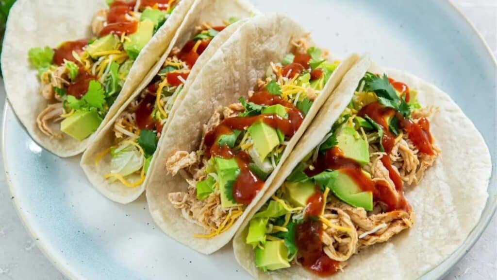 Three chicken tacos with avocado, cheese, cilantro, and salsa on a blue plate.