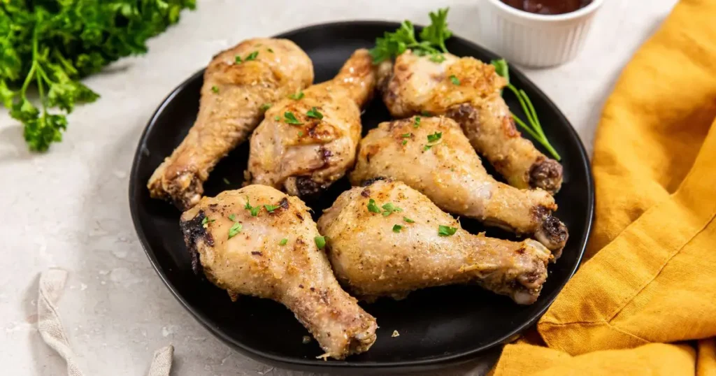 Plate of roasted chicken drumsticks garnished with parsley, accompanied by a bowl of sauce and a yellow napkin on a marble surface.