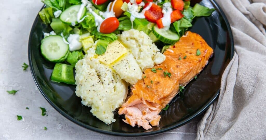 A well-balanced meal featuring baked salmon, mashed potatoes, and a leafy green salad with cherry tomatoes and cucumber on a black plate.