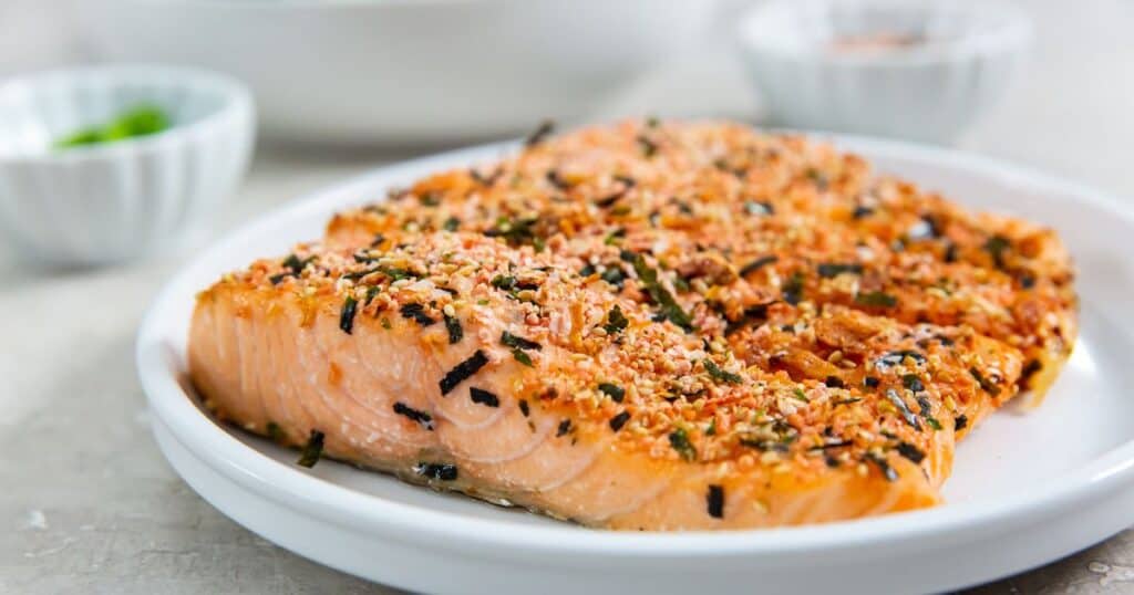 A cooked salmon fillet topped with herbs and spices on a white plate, with a small bowl of green herbs in the background.