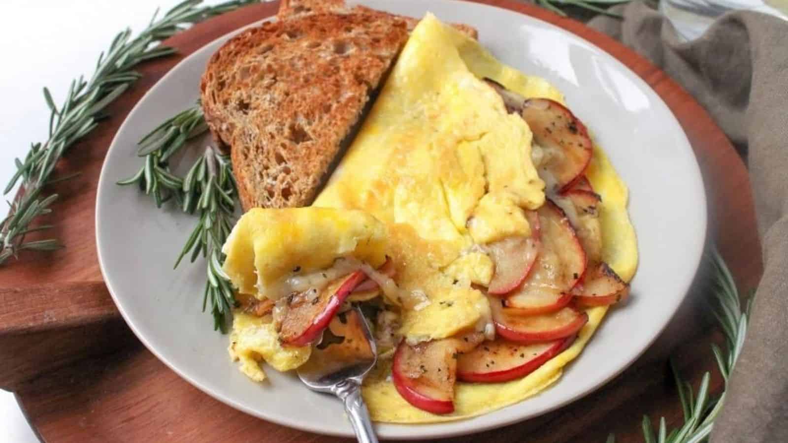 An apple white cheddar omelette with a slice of toast and a fork on a plate sitting on a wooden platter with fresh herbs surrounding it.