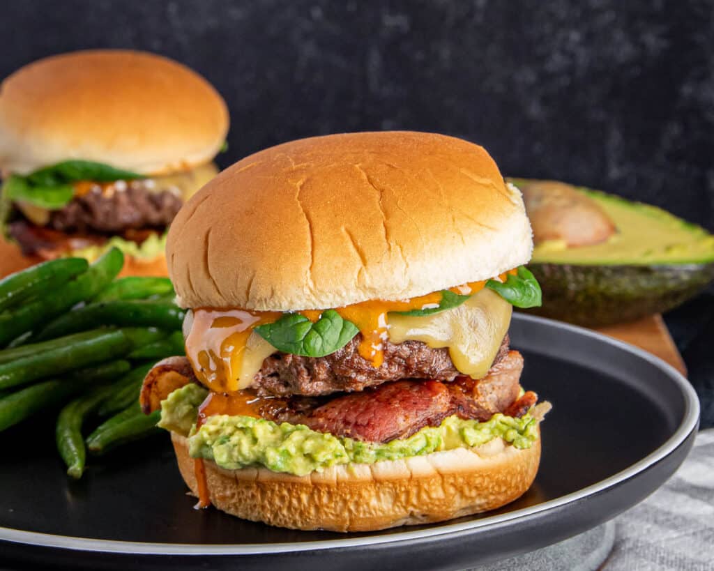 Juicy bacon cheeseburger with avocado and green beans on a black plate.