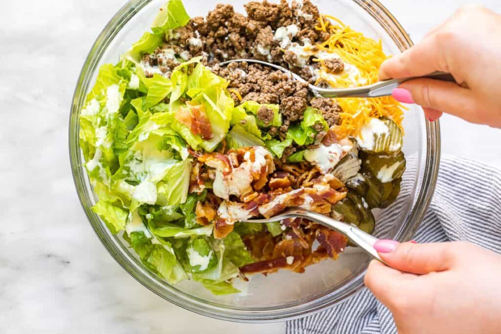 Two hands mix a salad with lettuce, ground beef, shredded cheese, bacon, and pickles in a glass bowl.