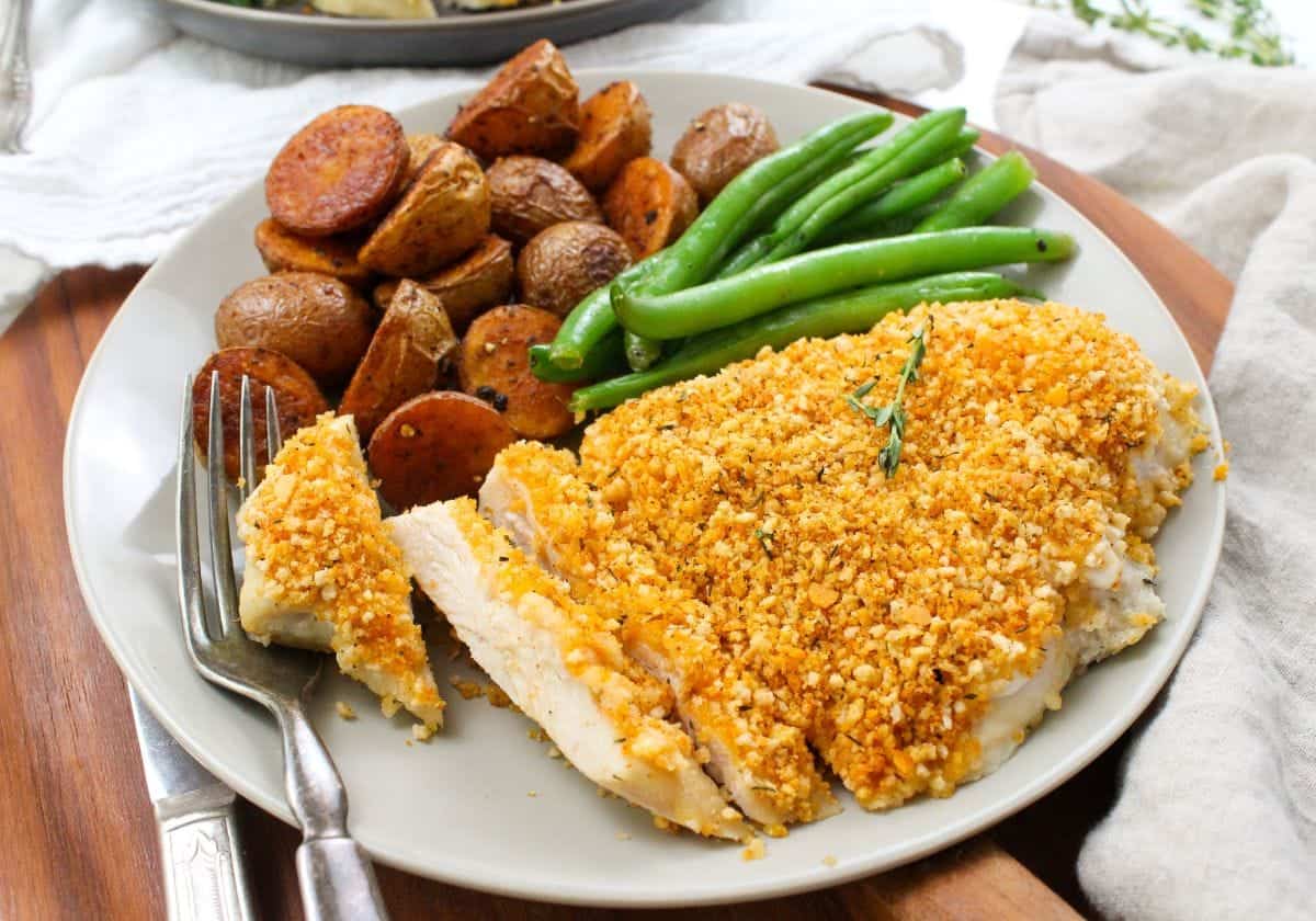 A sliced chicken breast coated in golden panko breadcrumbs on a plate next to green beans and roasted baby potatoes.
