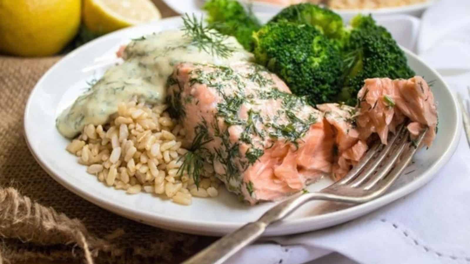 Salmon with creamy dill sauce on top next to brown rice and broccoli on a plate.