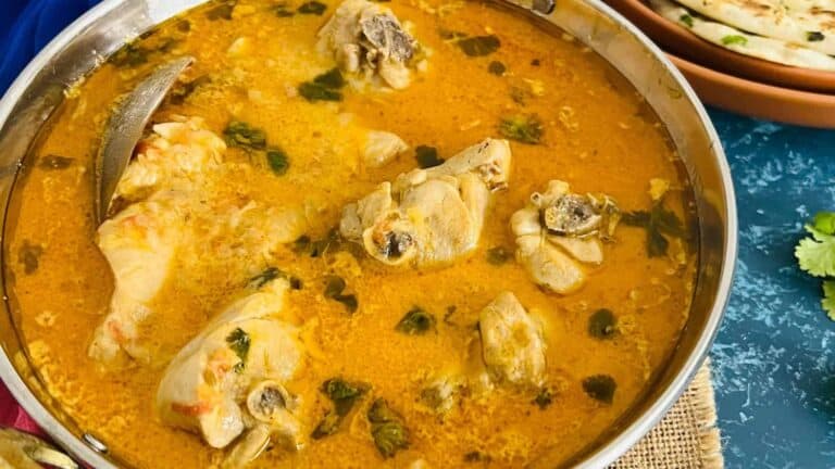 A bowl of traditional balti chicken garnished with cilantro, served next to naan bread on a blue tablecloth.