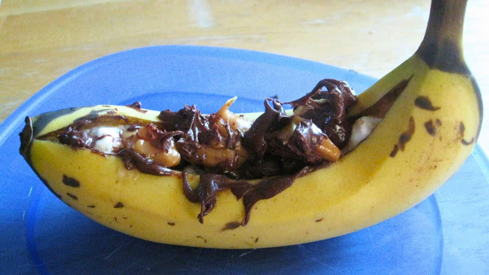 Image shows A ripe banana sliced open and filled with chocolate and caramel and marshmallow, served on a blue plate.