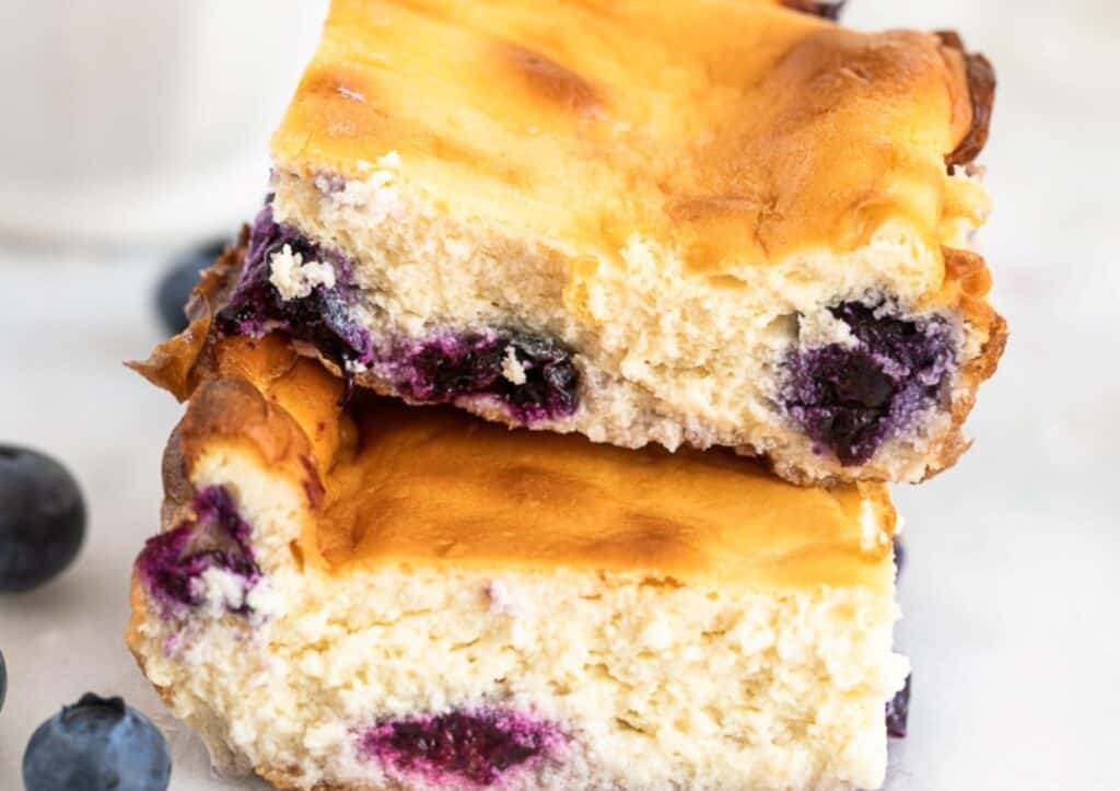 Close-up of blueberry cheesecake bars stacked on a plate, featuring golden brown tops and visible fruit pieces.