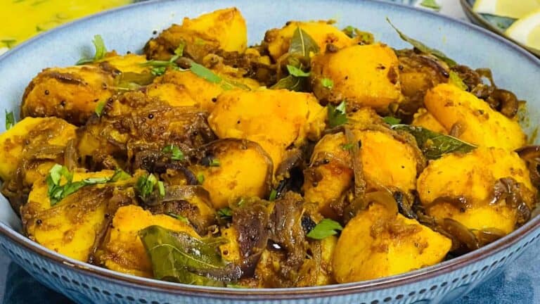 A bowl of Bombay Potatoes garnished with herbs, featuring golden potatoes and onions in a rich, spiced sauce.