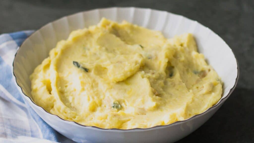 A bowl of brown butter garlic mashed potatoes on a table, next to a serving on a plate.