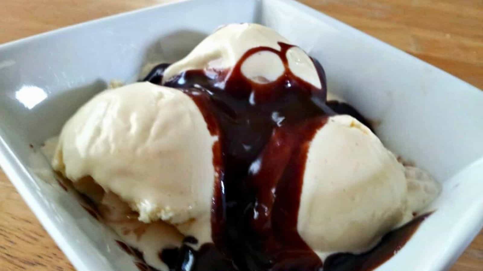 Image shows A bowl of vanilla ice cream topped with chocolate syrup on a wooden table.