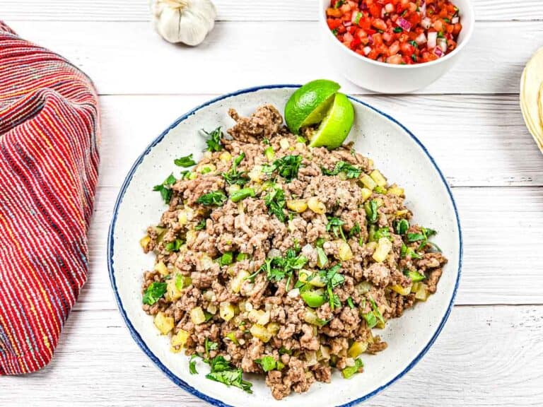 A plate of Mexican carne molida with lime wedges served on a wooden table with a striped cloth on the side.