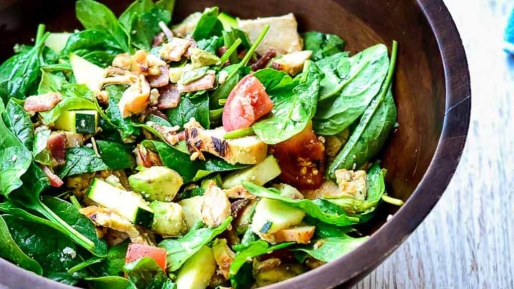 A fresh spinach salad with avocado, tomato, and bacon in a wooden bowl.