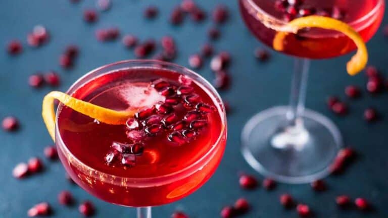 Two pomegranate cocktails garnished with orange peel, served in stemmed glasses on a blue surface with scattered pomegranate seeds.