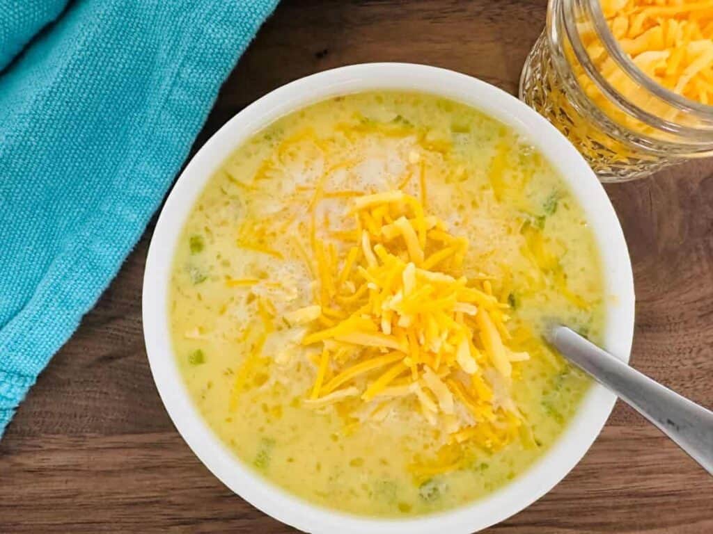 A bowl of creamy chicken soup topped with shredded cheese, accompanied by a jar of cheese and a blue napkin on a wooden surface.