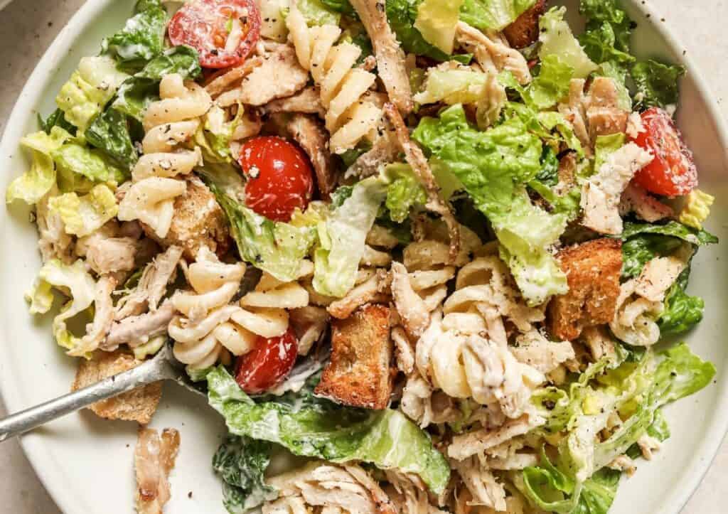 A close-up of a chicken Caesar salad with croutons, cherry tomatoes, and pasta, served in a white bowl with a fork.