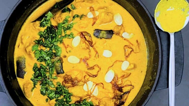 A bowl of Chicken Korma garnished with herbs and sliced almonds, served with a spoon on the side.