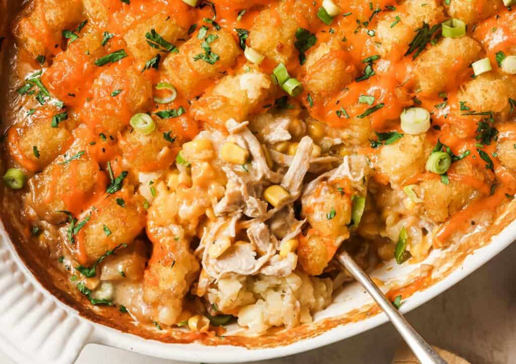 A casserole dish filled with tater tots, shredded chicken, and corn, topped with melted cheese and green onions.