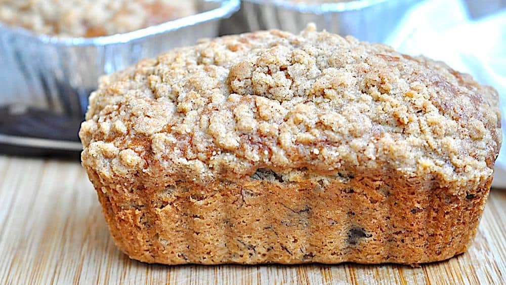 A close-up of a freshly baked loaf of banana bread with a golden-brown crust and a crumbly topping, set on a wooden surface.