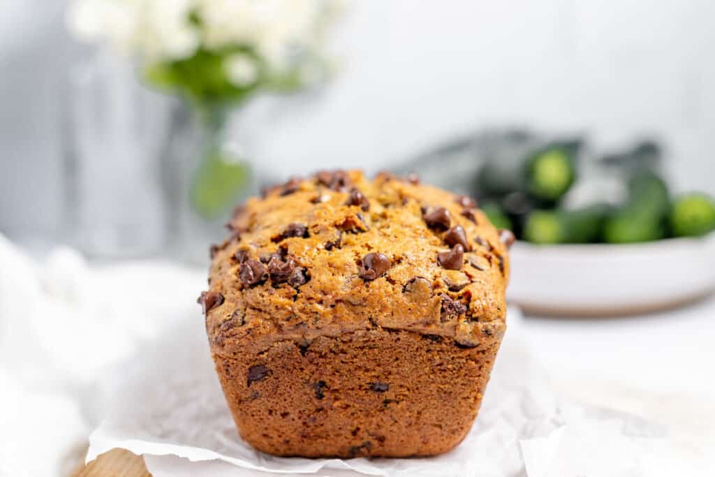A freshly baked chocolate chip muffin on a white paper with soft-focus flowers and vegetables in the background.