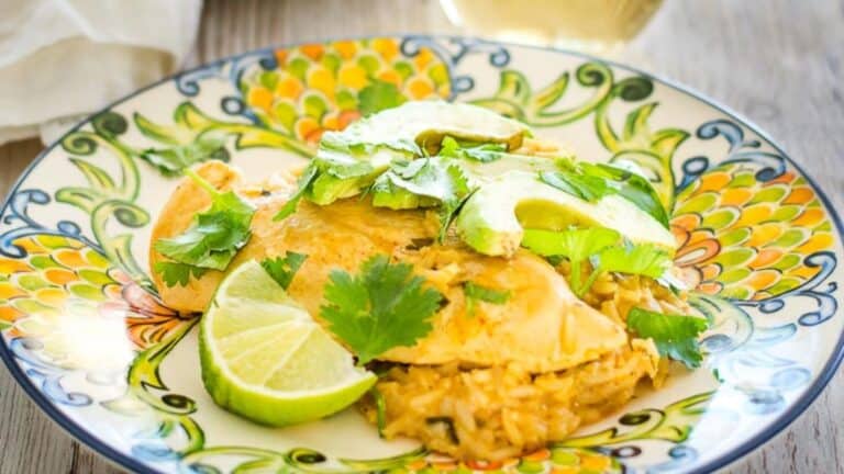 A plate of enchiladas topped with cheese, sour cream, avocado, cilantro, and a lime wedge on a decorative plate.