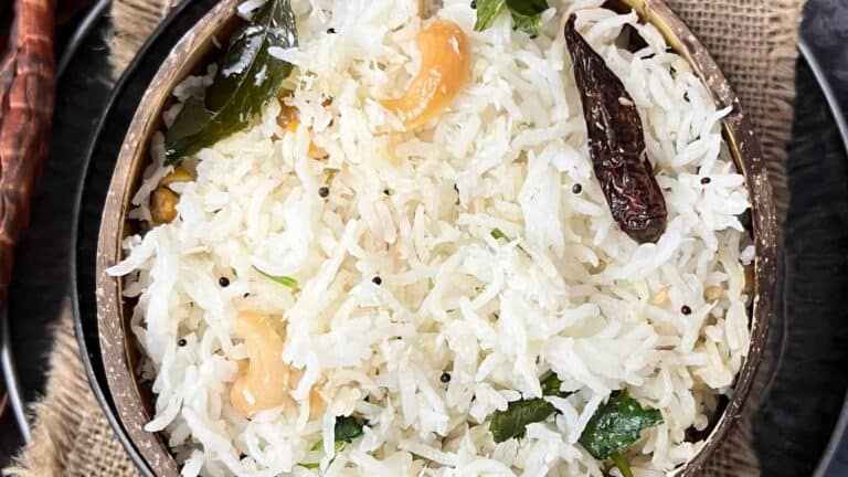 A bowl of cooked white rice garnished with cashews, curry leaves, and dried red chilies, displayed on a rustic table setting.