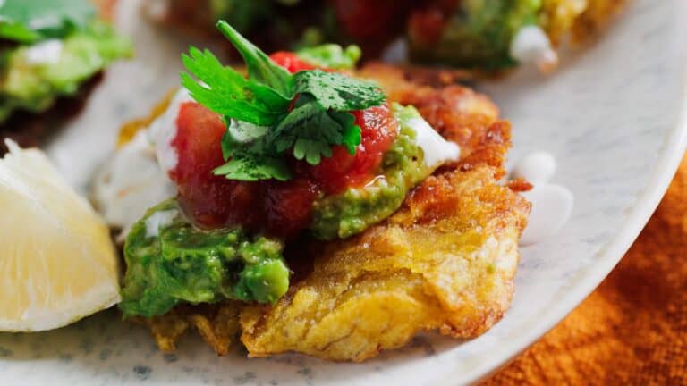 A plate of fried green plantains topped with guacamole, salsa, and a dollop of cream, garnished with cilantro and a lemon wedge on the side.