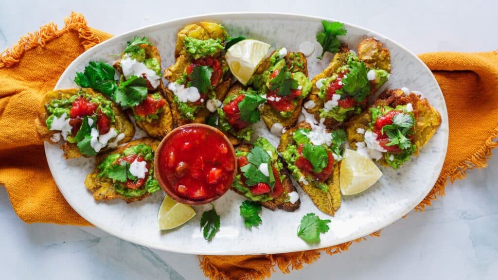 A plate of crispy tostadas topped with salsa, guacamole, and crumbled cheese, garnished with cilantro and lime wedges, alongside a bowl of red salsa.