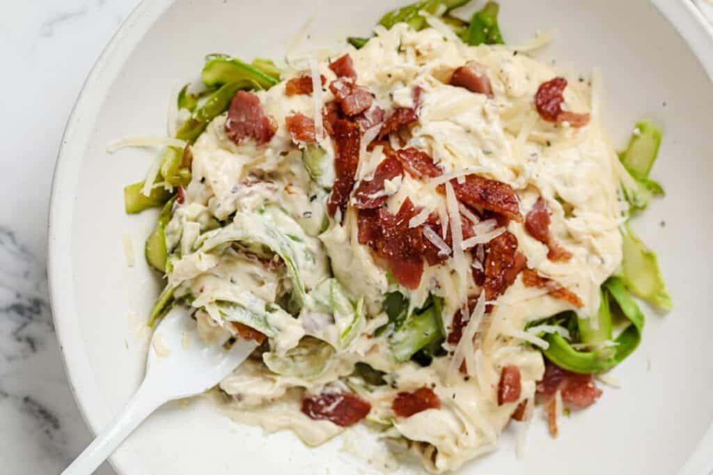 A bowl of zucchini noodles topped with creamy sauce and crispy bacon pieces, served with a spoon on a marble surface.