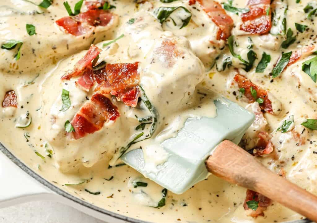 Creamy chicken dish garnished with bacon and herbs in a skillet with a spatula.