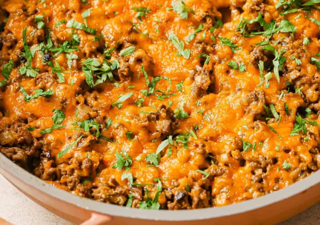 A close-up of a skillet filled with cooked ground meat topped with melted cheese and fresh parsley.