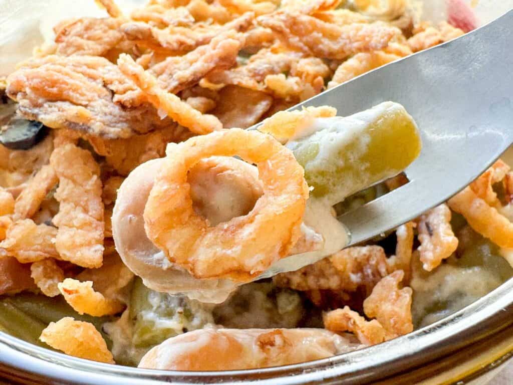 A close-up of a green bean casserole topped with crispy fried onions, with a spoon lifting some from the dish as someone asks for seconds.
