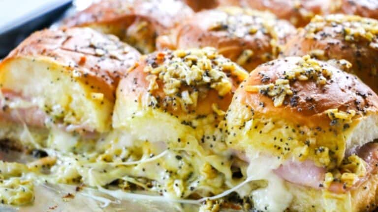 Baked sliders with melted cheese and ham on poppy seed-topped buns in a baking dish.