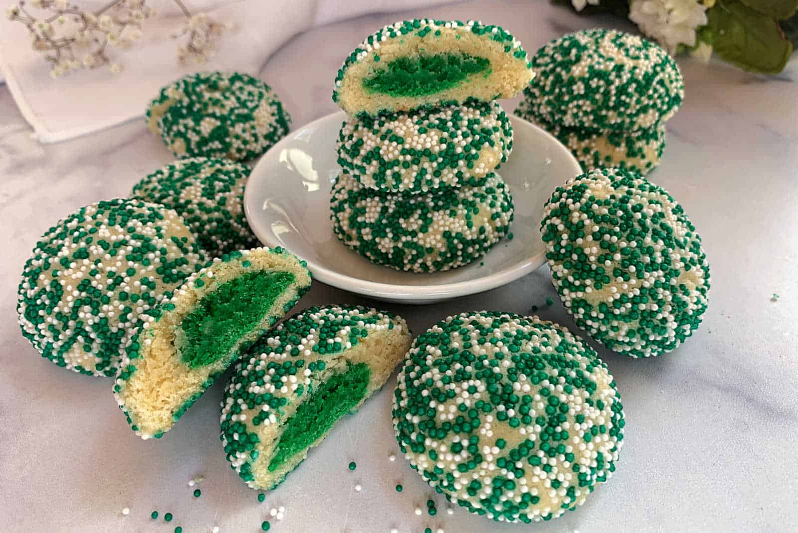 A plate of green and white sprinkled cookies, some cut in half to show green centers, next to a cup of tea on a table with a lacy placemat.