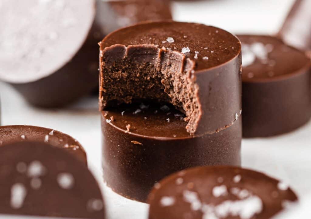 Close-up of a bitten chocolate dessert sprinkled with sea salt, surrounded by whole chocolates on a white surface.