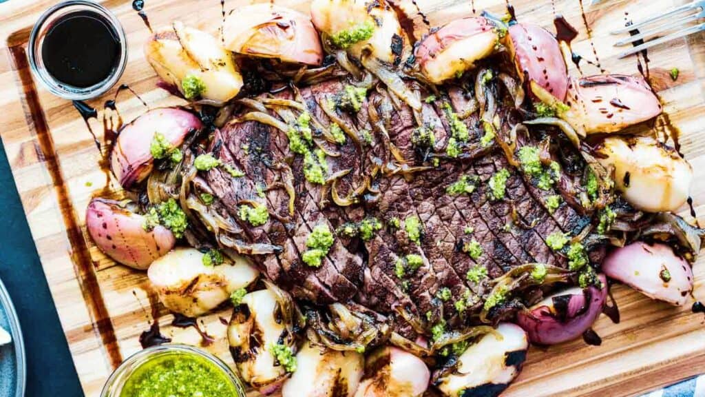 Grilled steak topped with chimichurri sauce, surrounded by roasted onions and potatoes on a wooden cutting board.