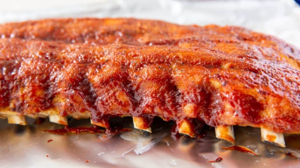Close-up of glazed barbecued ribs on aluminum foil, highlighting the shiny, sticky sauce and visible meat texture.