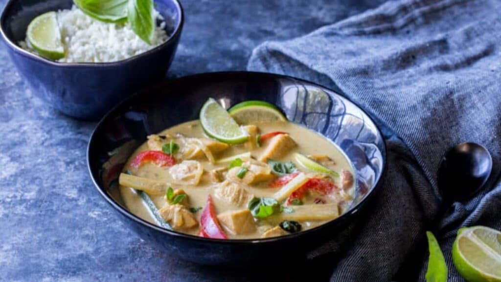 Bowl of thai chicken curry with slices of red bell pepper and lime, served with a side of rice, on a slate background.