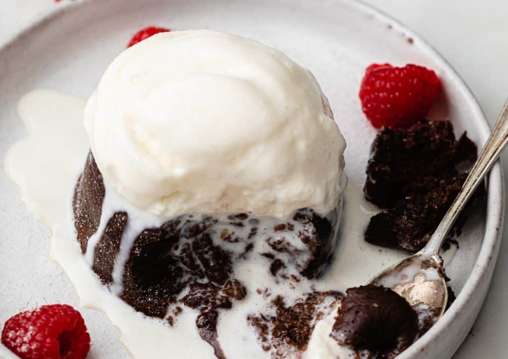 A scoop of vanilla ice cream melting over a warm chocolate lava cake, garnished with fresh raspberries on a white plate.