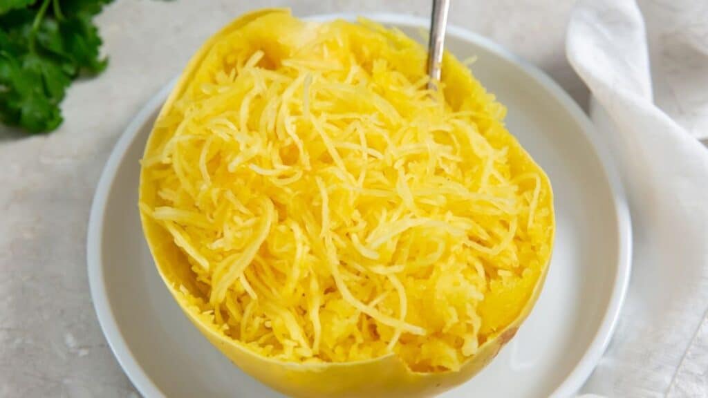 Cooked spaghetti squash with strands separated, served in its half shell on a white plate.