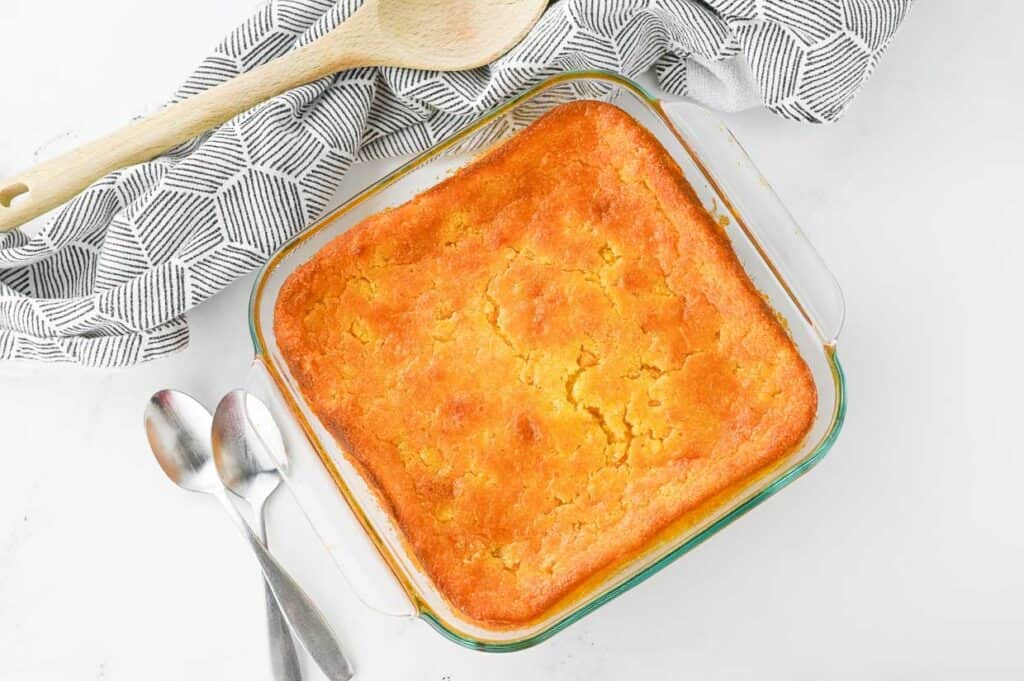 A freshly baked cornbread in a glass dish with a cloth and wooden spoon beside it, on a white surface, tempting everyone to start asking for seconds.