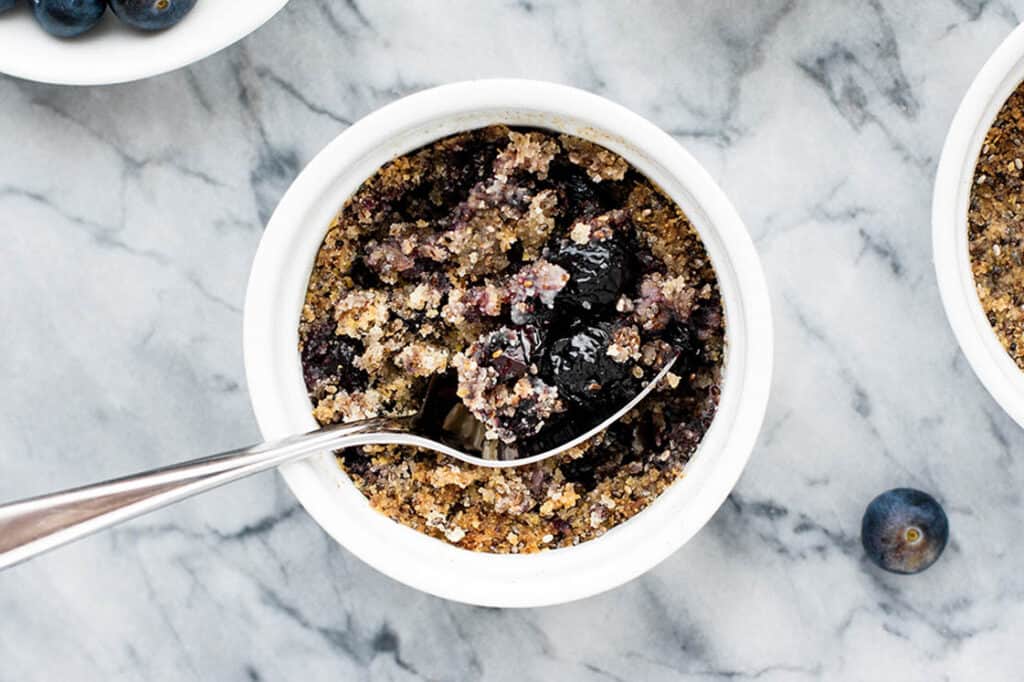 A bowl of blueberry crumble with a spoon on a marble countertop, accompanied by fresh blueberries and another bowl in the background.