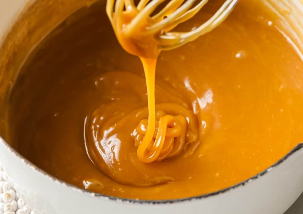 Whisk lifting thick, smooth caramel sauce from a white bowl.