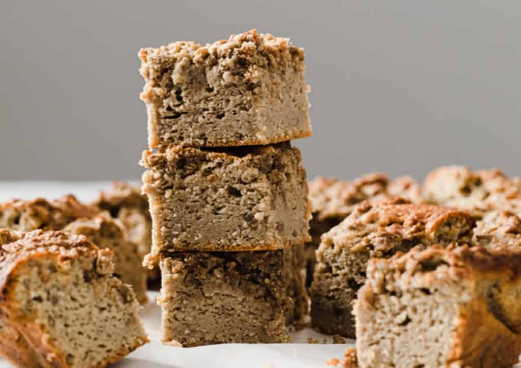 Stack of dense, freshly-baked coffee cake slices with a textured, crumbly crust, set against a soft-focus grey background.