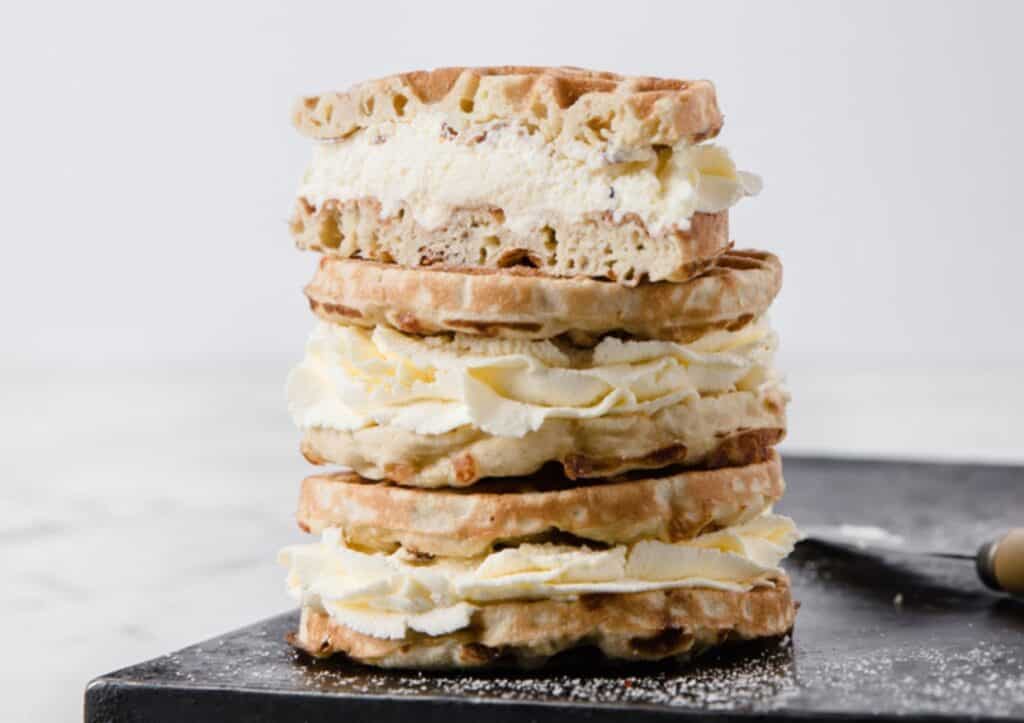 A stack of crisp waffles with layers of whipped cream on a dark slate board, with a knife to the side.