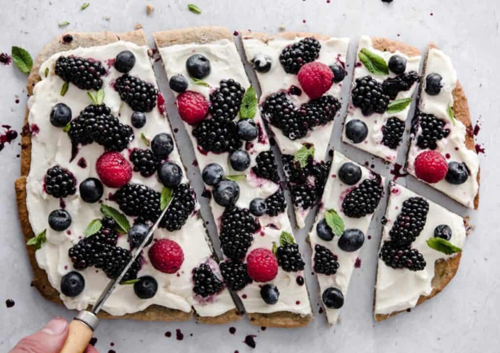 A dessert pizza with a cream cheese topping, garnished with blackberries, blueberries, raspberries, and fresh mint leaves, sliced and served on a white surface.