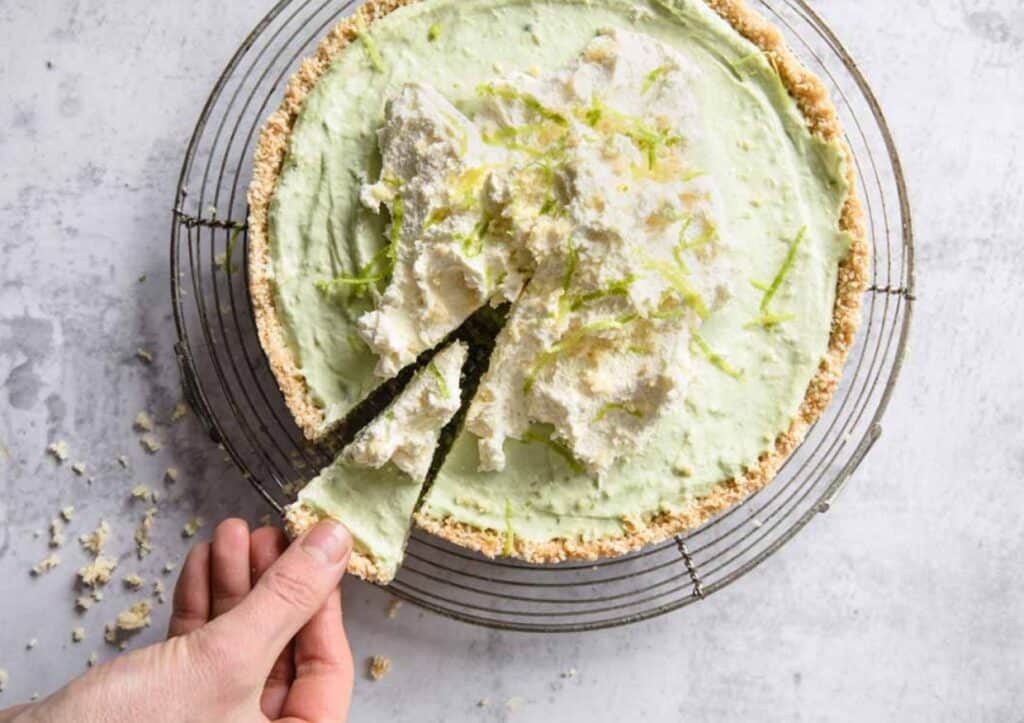 A hand serving a slice of lime-flavored pie with a crumbly crust, topped with whipped cream and zest, on a wire cooling rack.